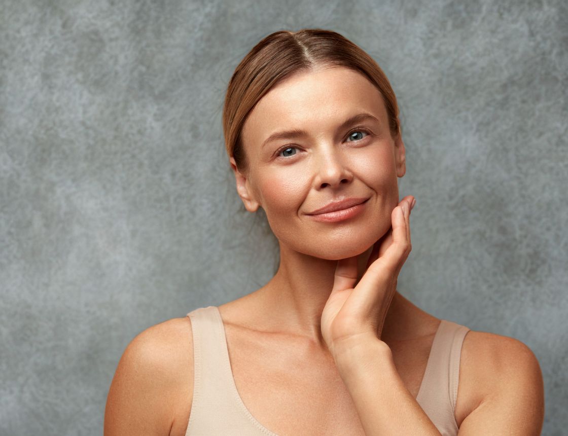Close-up studio portrait of a person with clear, healthy-looking skin, wearing a neutral beige top and gently touching the side of the face against a soft grey background.